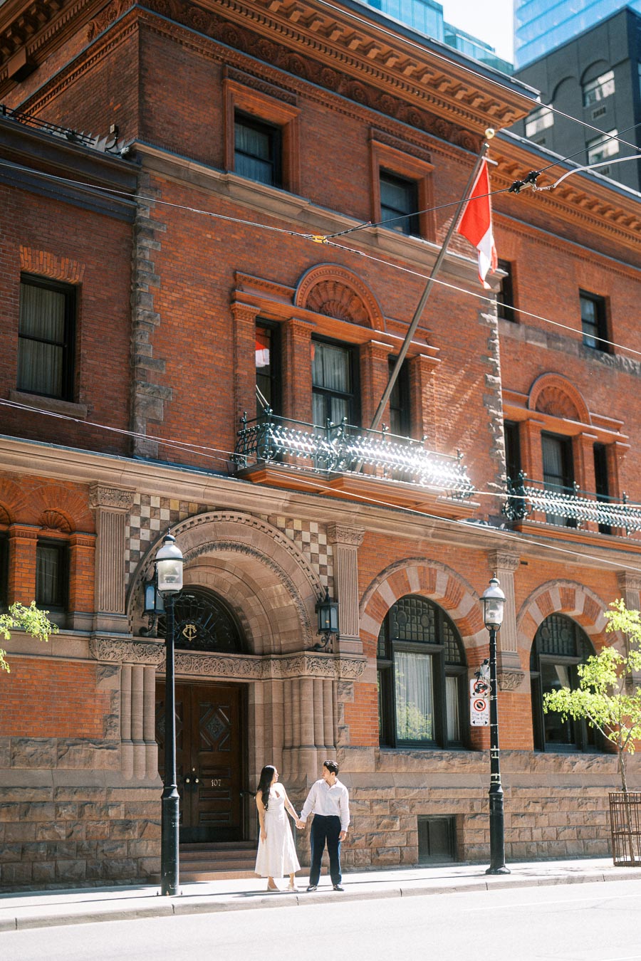 Cassie and Gabriel standing and holding hands on a city sidewalk in front of a large historic brick building in downtown Toronto.