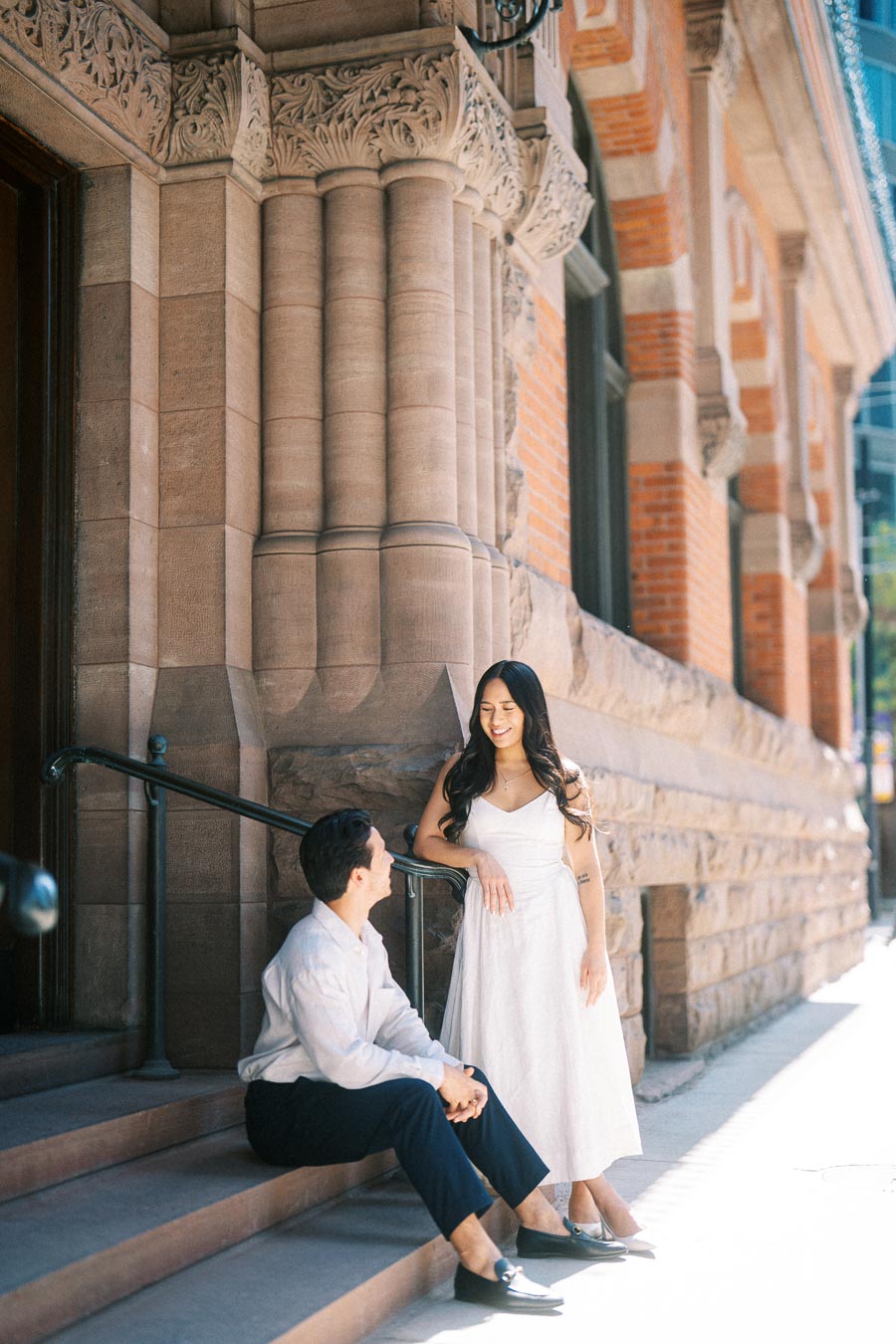 Cassie and Gabriel during their engagement session, with Cassie standing and Gabriel sitting on stone steps outside a historic building in downtown Toronto.