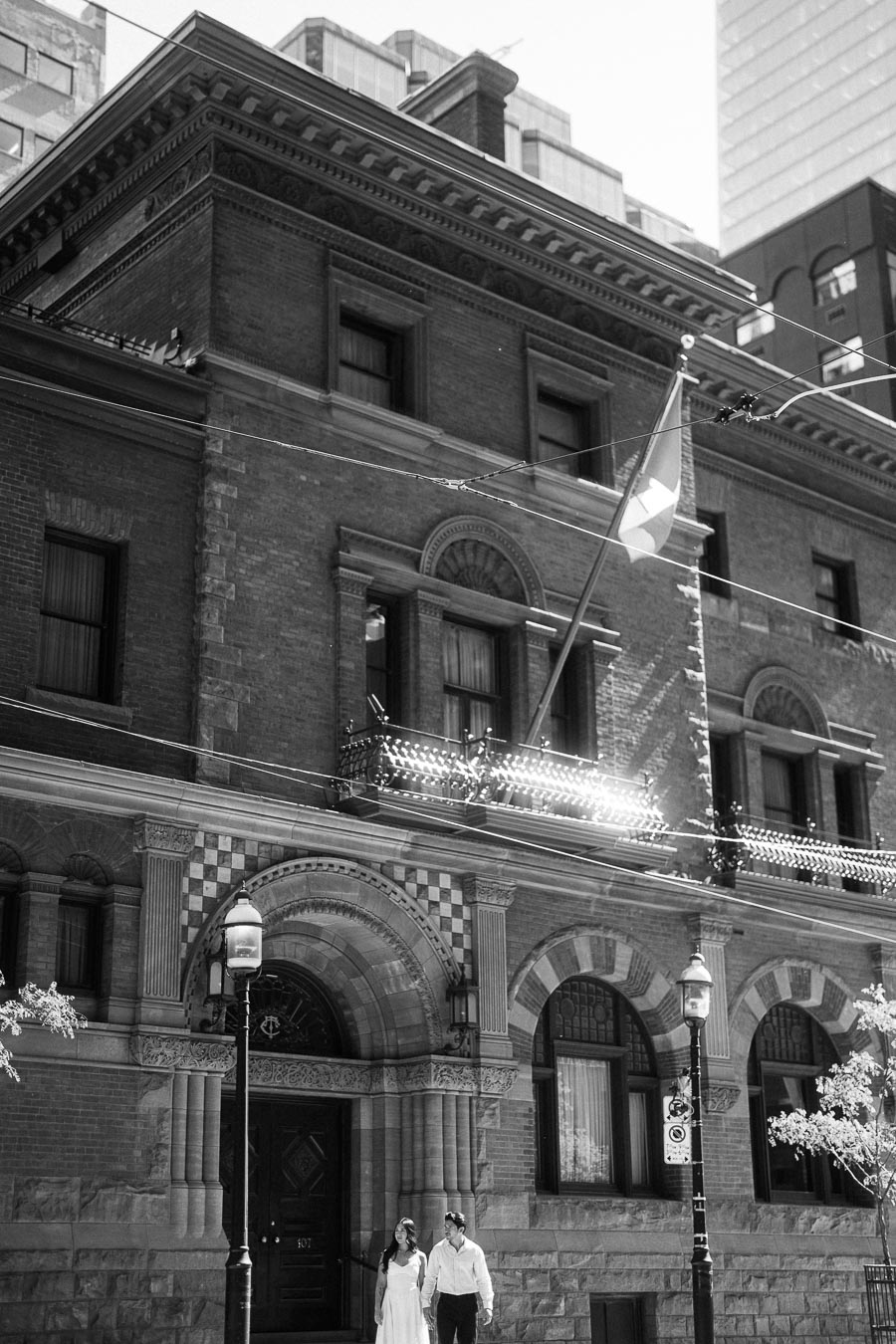 Cassie and Gabriel walk together outside a historic brick building with arched windows and a flag in downtown Toronto.