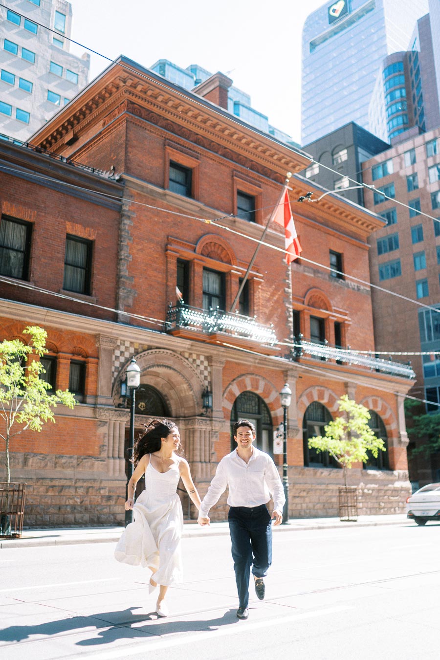 Cassie and Gabriel run hand in hand in front of a historic brick building with a Canadian flag in downtown Toronto.