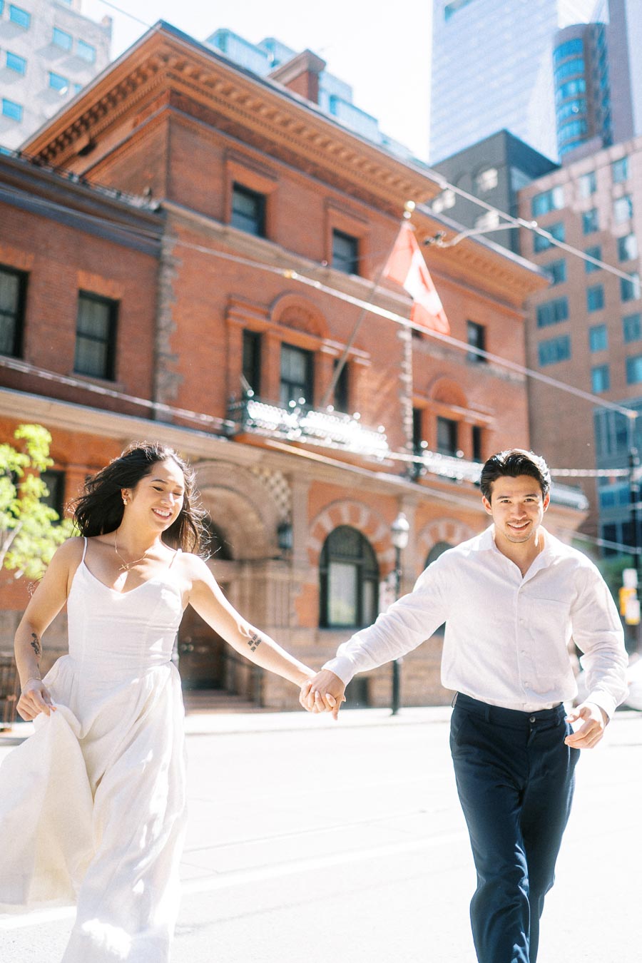 Cassie and Gabriel holding hands and running on a sunlit downtown Toronto street with a historic brick building and Canadian flag in the background.