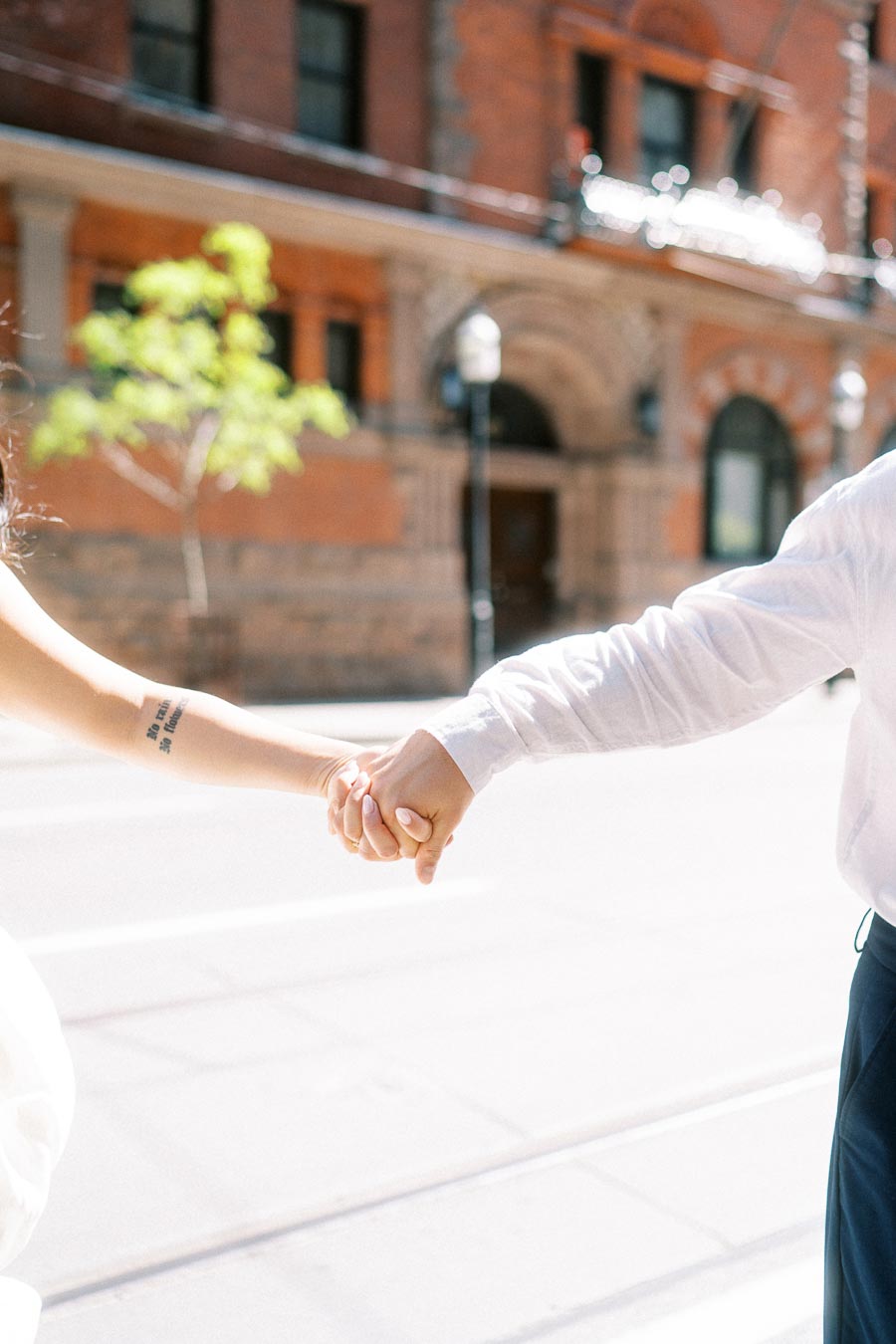 Close-up of Cassie and Gabriel holding hands on a bright city street in downtown Toronto, with a historic brick building in the background.