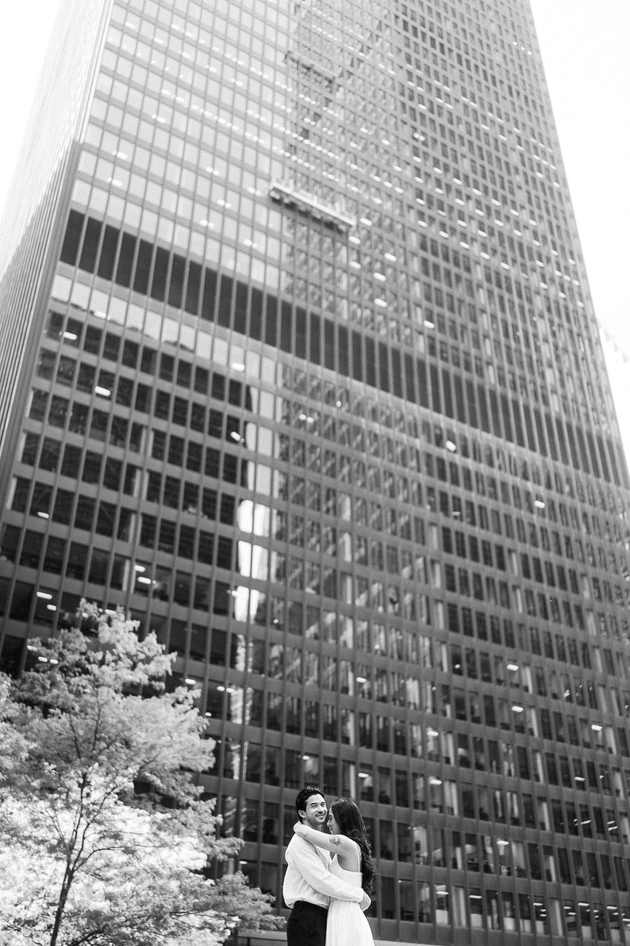 Cassie and Gabriel embracing and smiling in front of a tall glass office building in downtown Toronto during their engagement session.