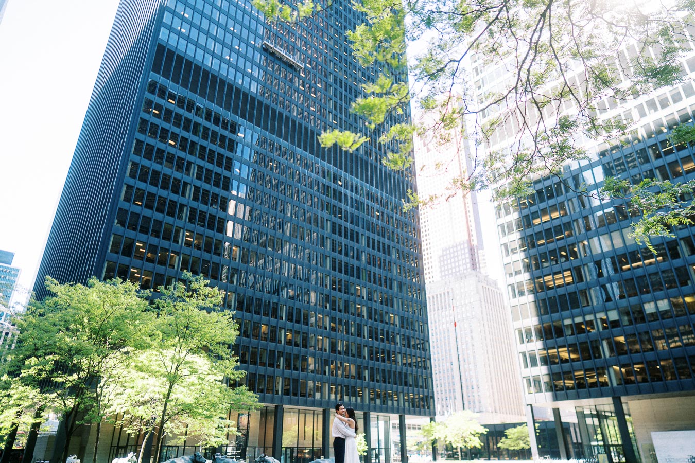 Cassie and Gabriel hugging in the center of a plaza, framed by tall glass office buildings and leafy trees in downtown Toronto.