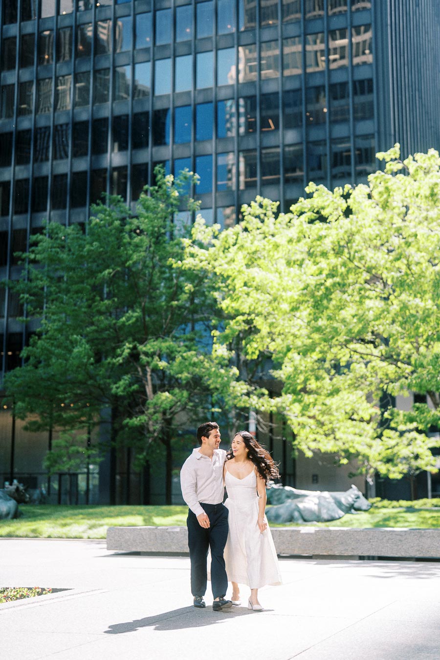Cassie and Gabriel walking arm in arm on a sunny day in downtown Toronto, with trees and glass office buildings in the background.