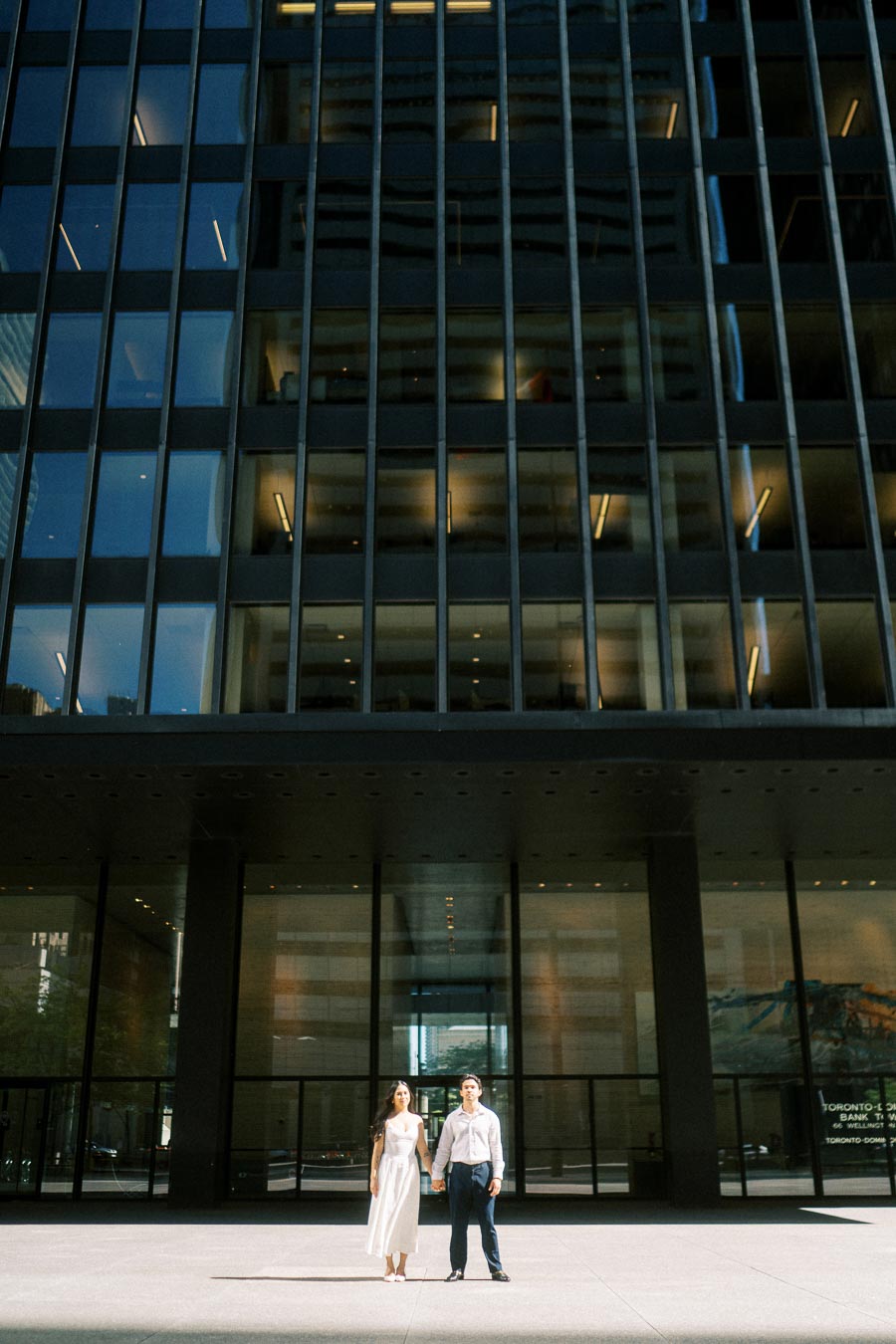 Cassie and Gabriel holding hands in front of a tall glass office tower in downtown Toronto during their engagement photoshoot.