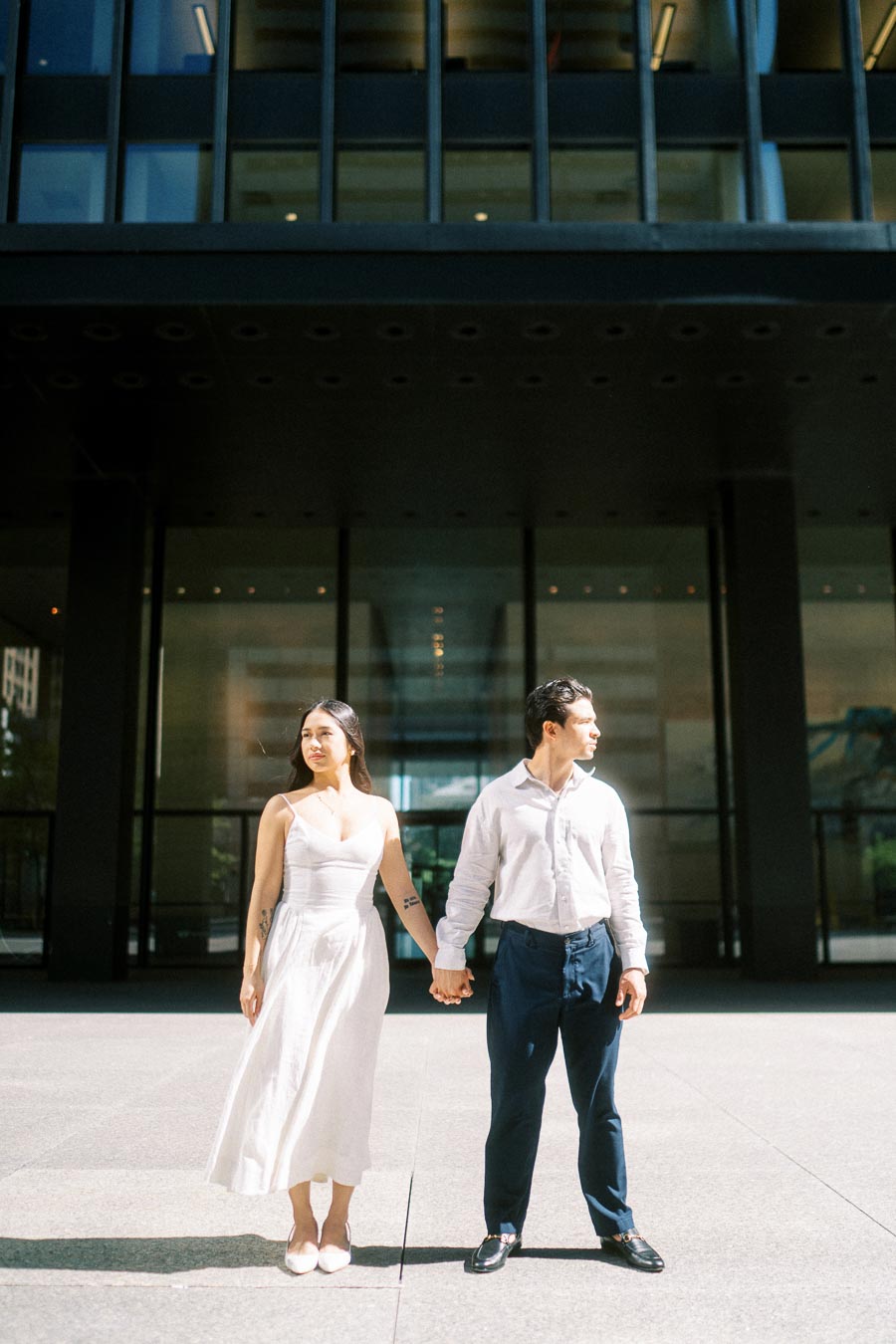Cassie and Gabriel stand holding hands in sunlight in front of a modern glass office building in downtown Toronto.