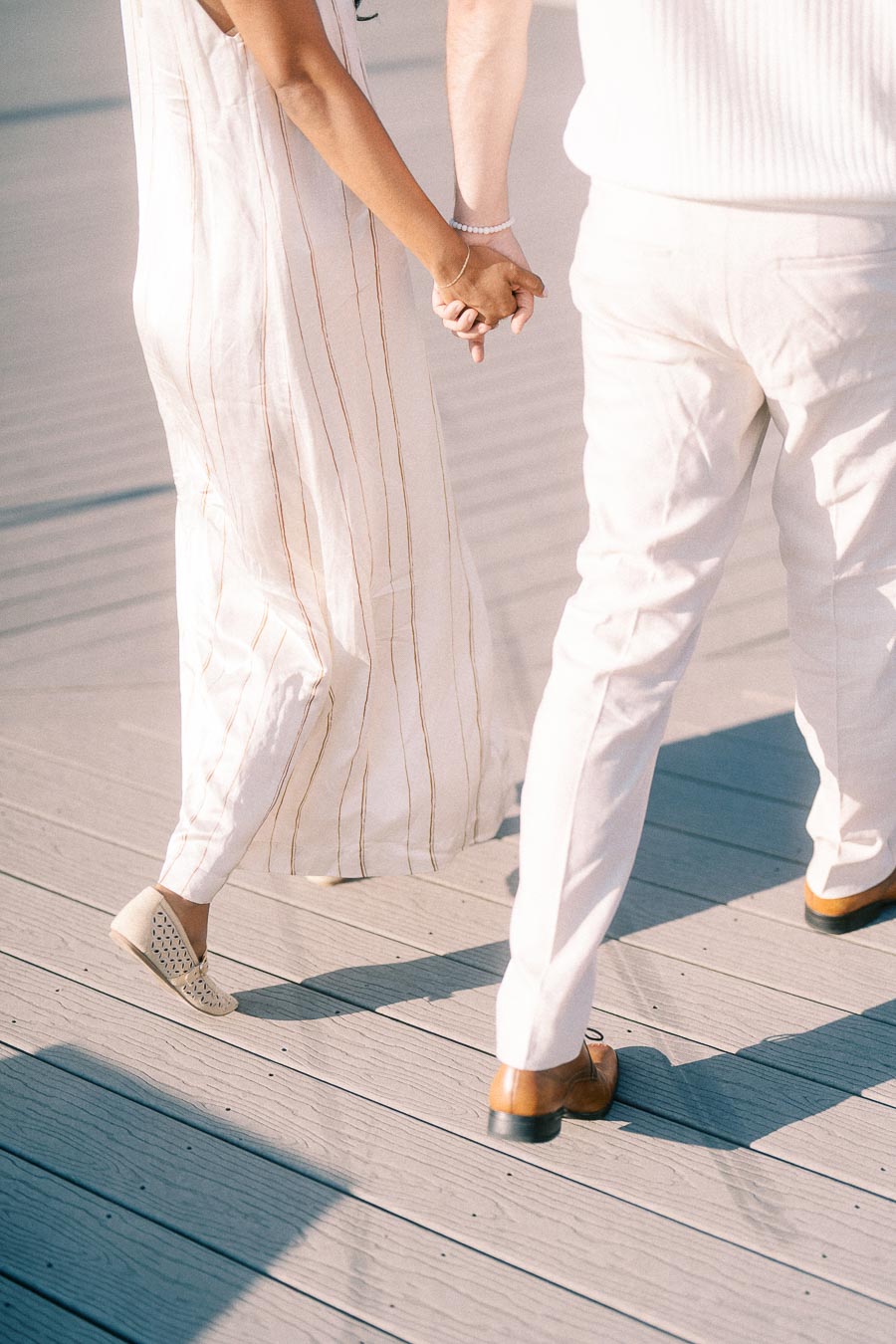 Couple holding hands while walking on a sunlit marina dock during an engagement session.