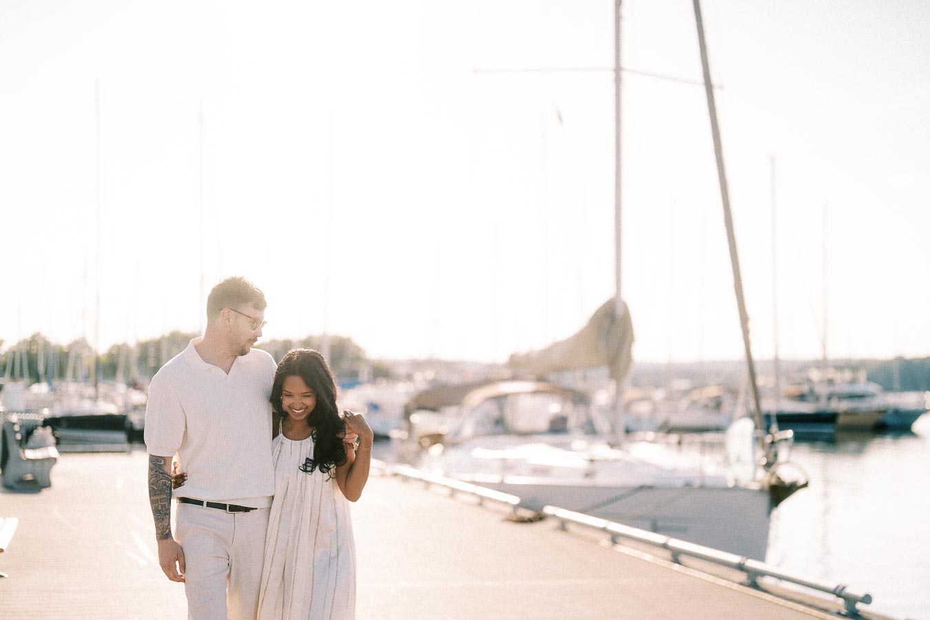 Amber and Devin walking together on a marina dock beside sailboats at golden hour.