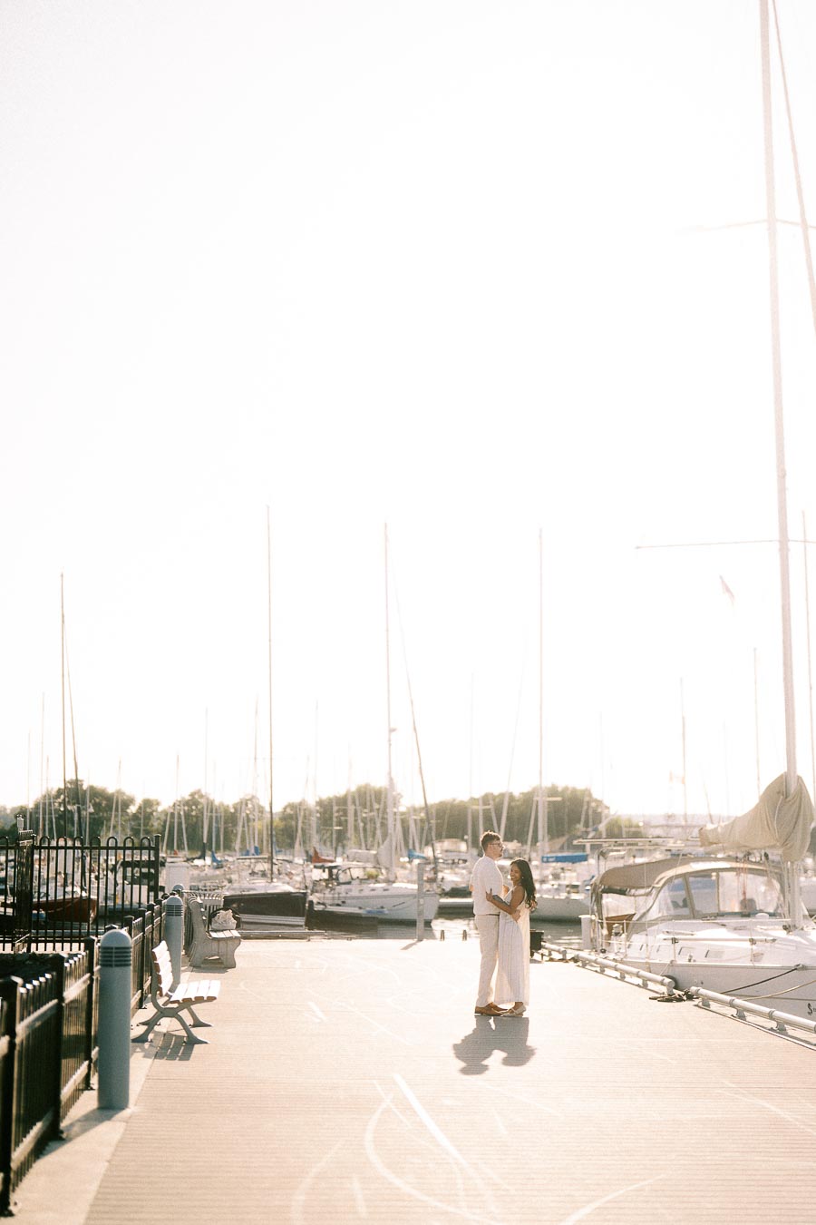 Amber and Devin embrace on a sunlit marina dock beside sailboats in Hamilton, Ontario.