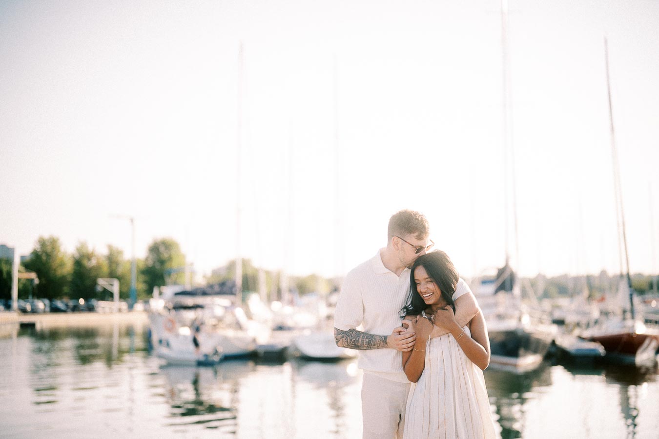 Amber and Devin embrace by sailboats at a Hamilton marina during golden hour.