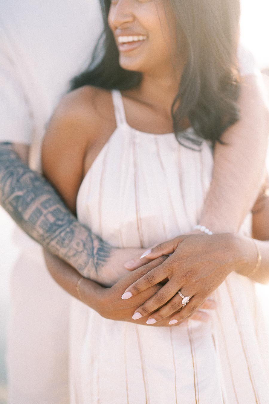 Smiling woman with engagement ring embraced by her partner in warm golden-hour light.