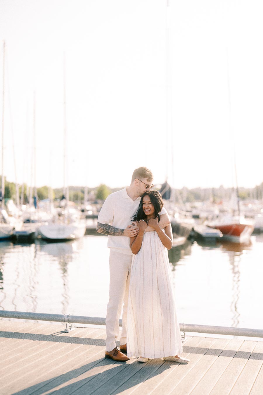 Amber and Devin embracing on a marina dock with sailboats behind them at golden hour.