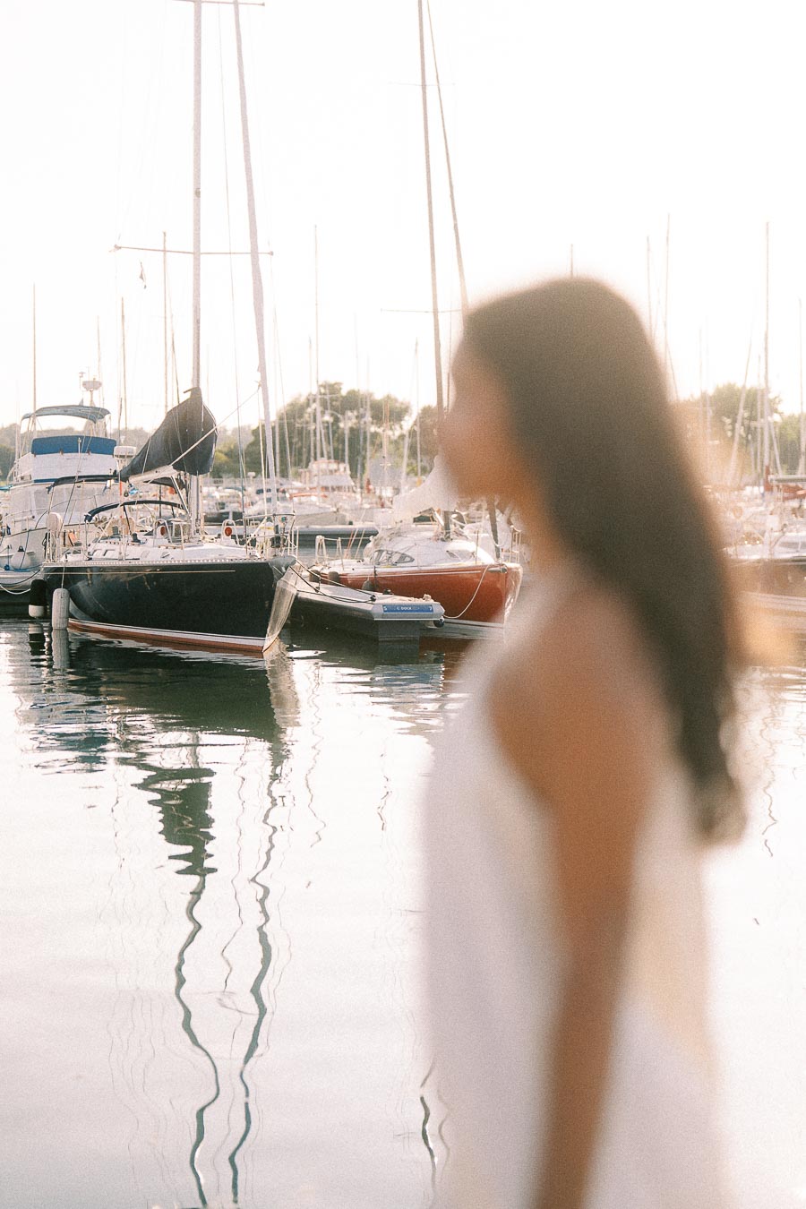 Blurred woman in white dress beside sailboats at a sunny Hamilton marina.