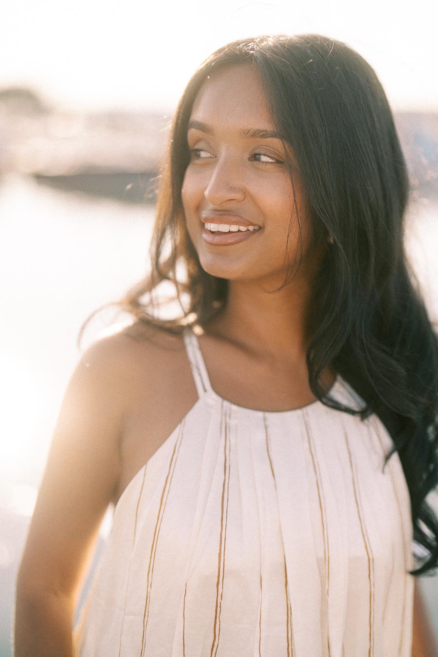 Smiling woman in a white dress backlit by golden hour sun at a waterfront marina.