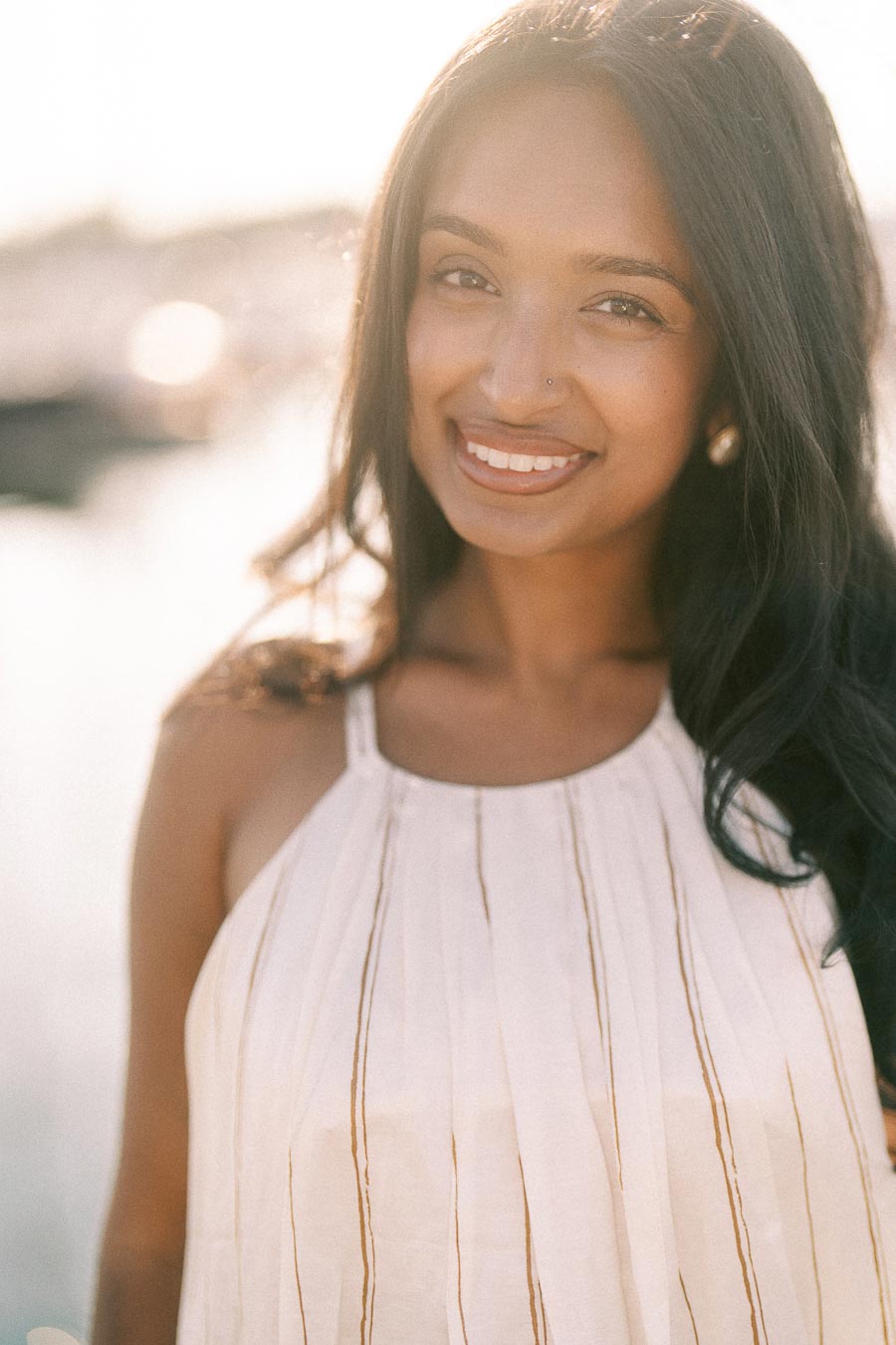 Smiling woman in a white dress photographed in warm light by the marina waterfront.
