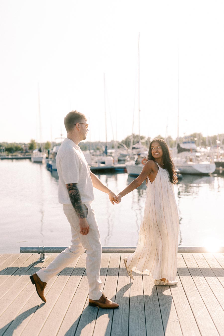 Amber and Devin holding hands on a marina dock with sailboats behind them in Hamilton, Ontario.