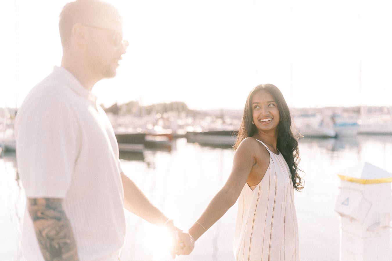 Amber and Devin holding hands by sailboats at a sunny Hamilton marina engagement session.