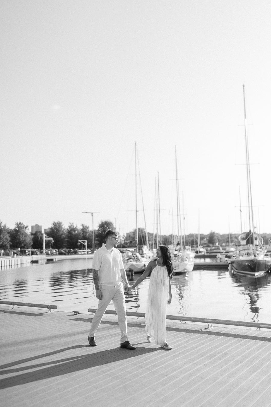 Amber and Devin hold hands on a marina dock with sailboats behind them in Hamilton, Ontario.
