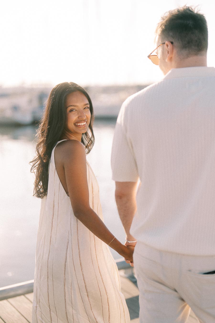 Amber smiles back while holding Devin’s hand on a marina dock at golden hour.