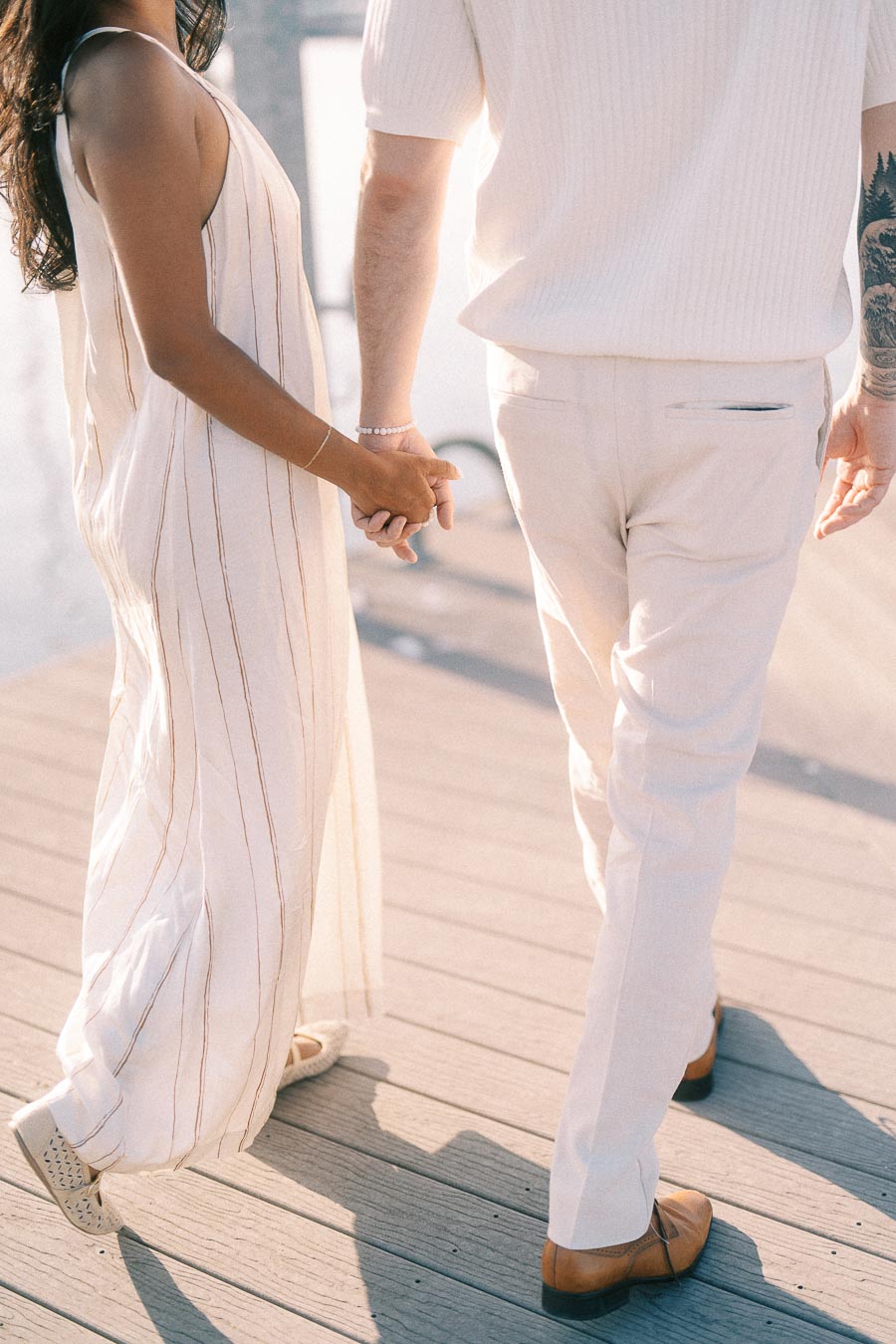 Couple holding hands while walking on a sunlit marina dock during an engagement session.