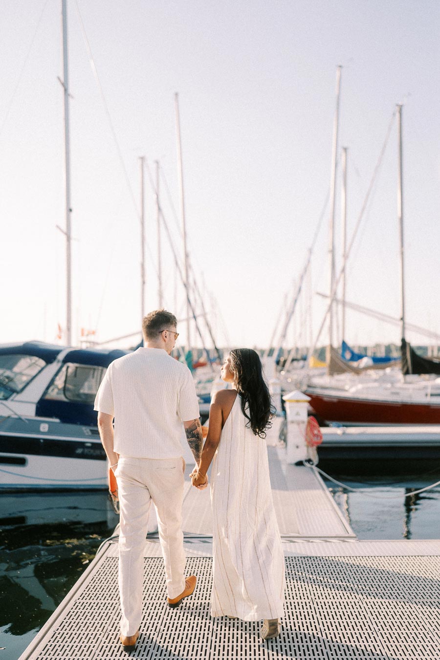 Amber and Devin hold hands while walking along a marina dock with sailboats at golden hour.