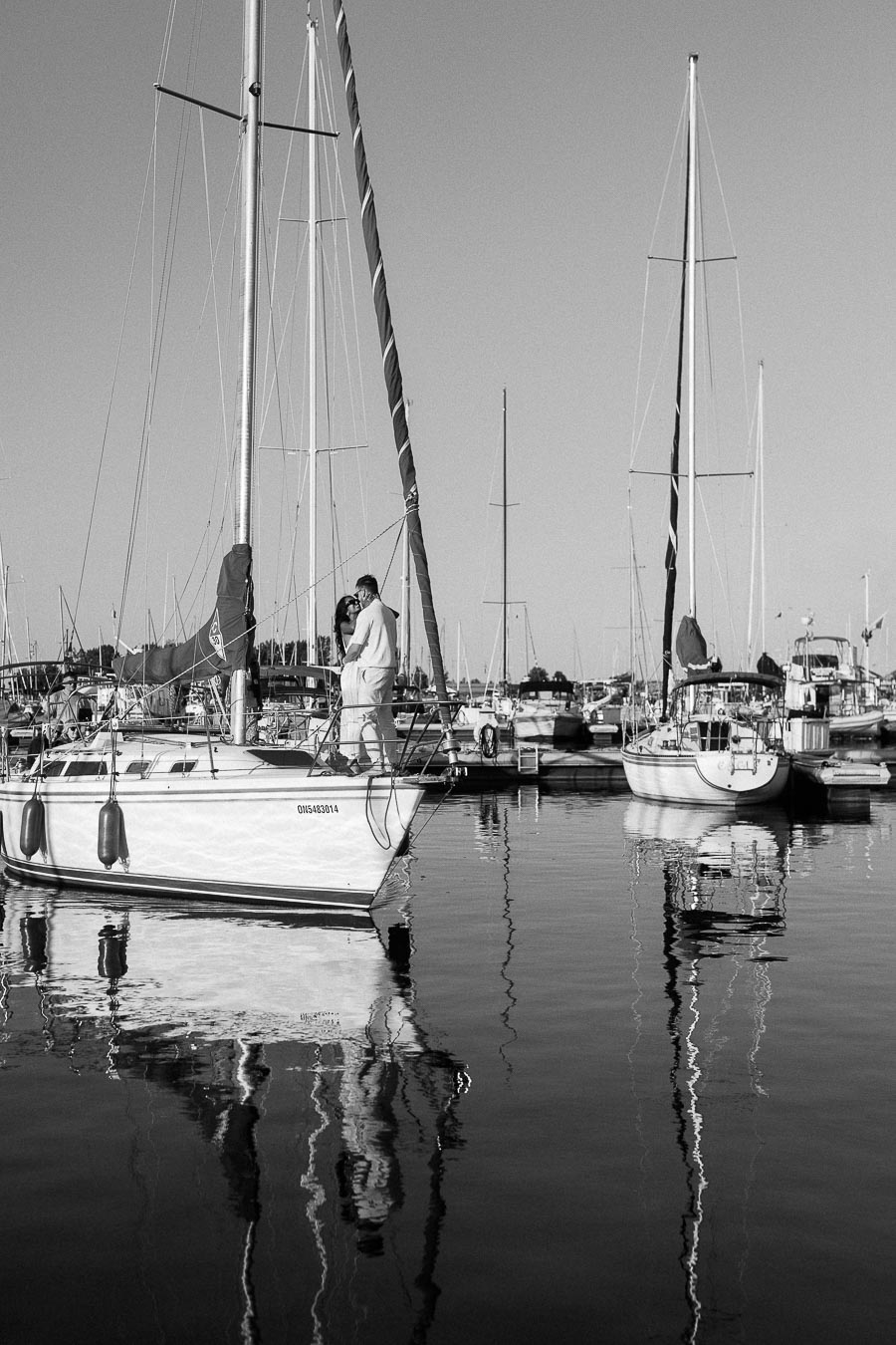 Couple embraces on a sailboat in a marina with masts reflected in calm water.