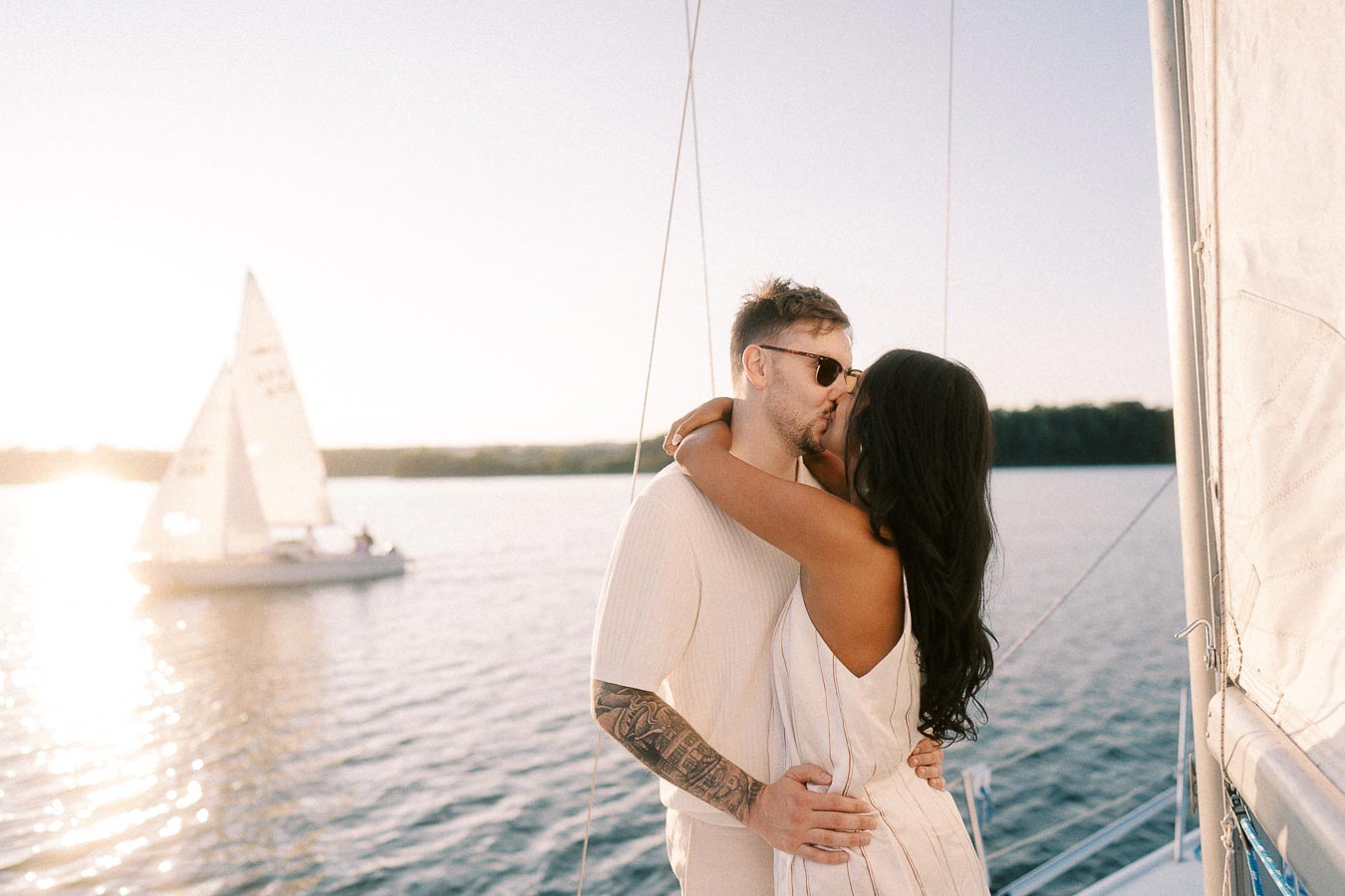 Amber and Devin kissing on a sailboat with another sailboat in the sunset background.