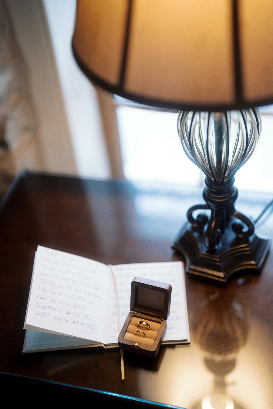 Open ring box with two rings beside a handwritten notebook and table lamp.