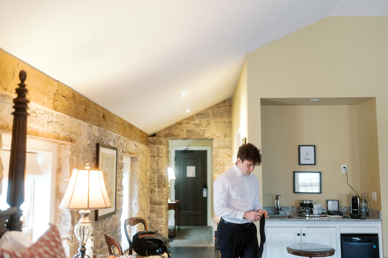 Man in a white shirt adjusting a black tie in a warm stone-walled room