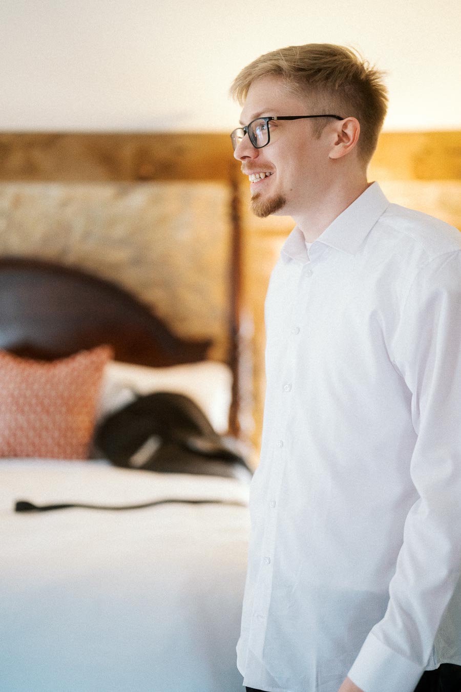 Man in glasses and a white shirt smiling beside a bed indoors