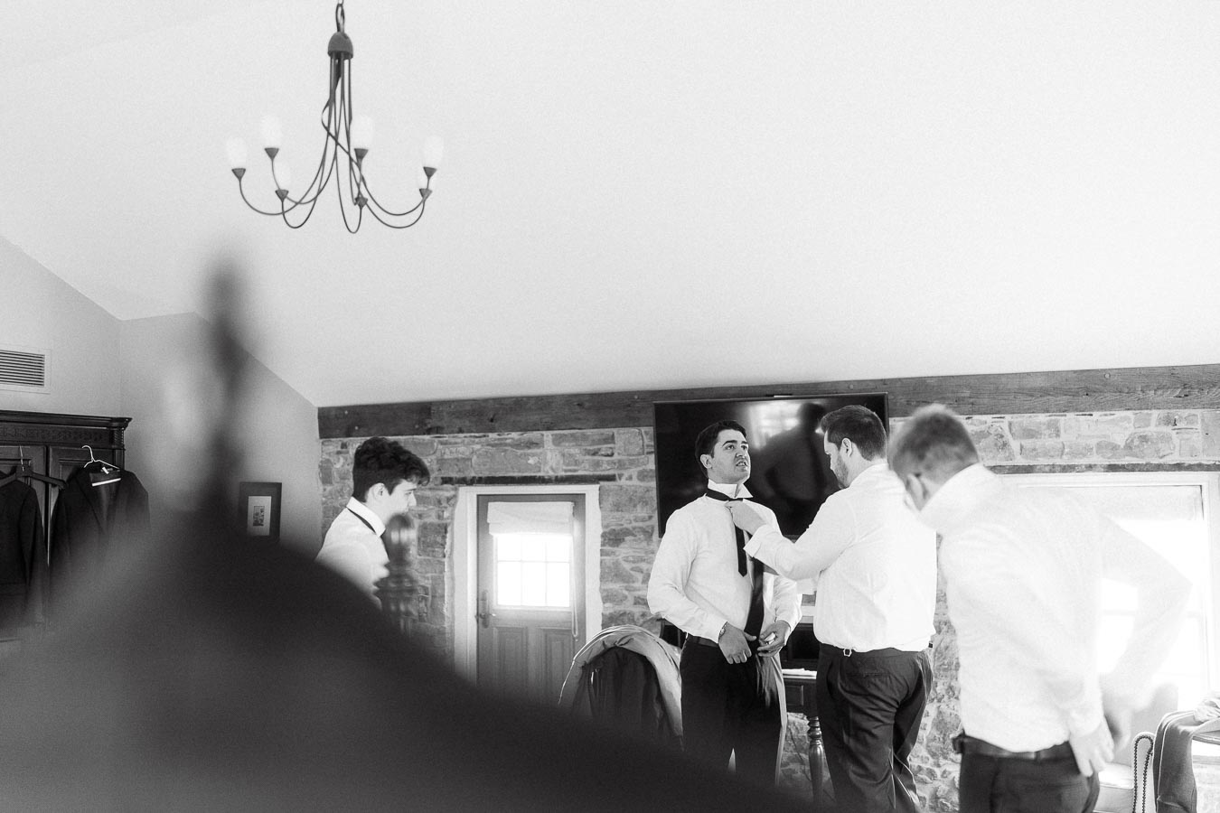 Men helping a groom adjust his tie in a stone-walled room.