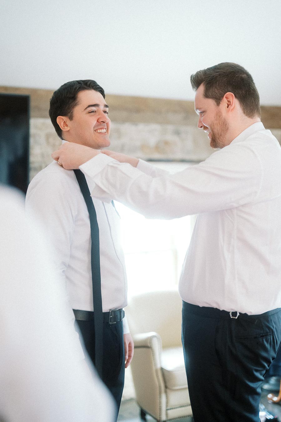 Two men in dress shirts smile as one adjusts the other's collar.