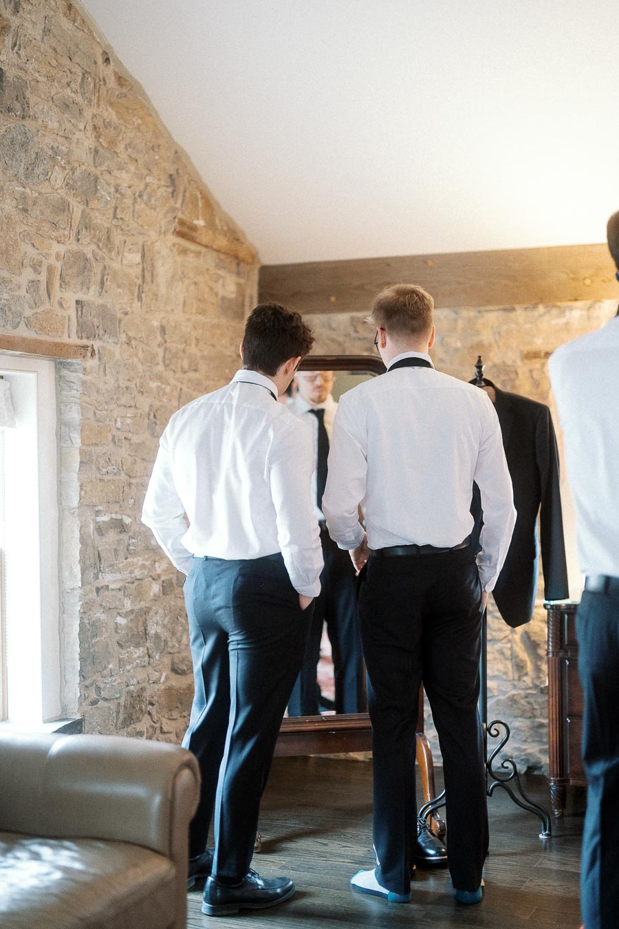 Two men in dress shirts adjust formalwear in front of a mirror in a stone-walled room.