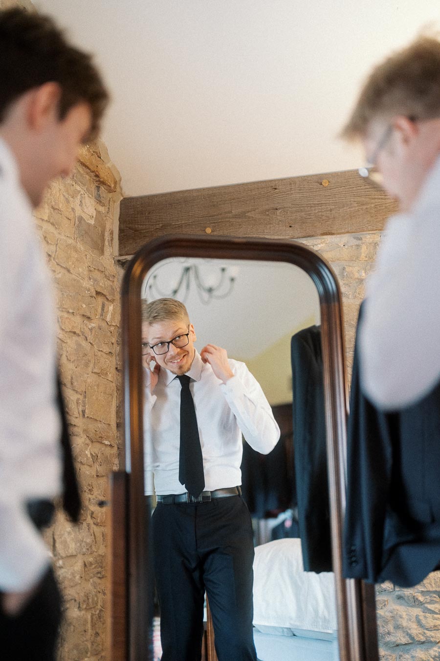Man adjusting his shirt collar in a mirror while another man stands nearby.
