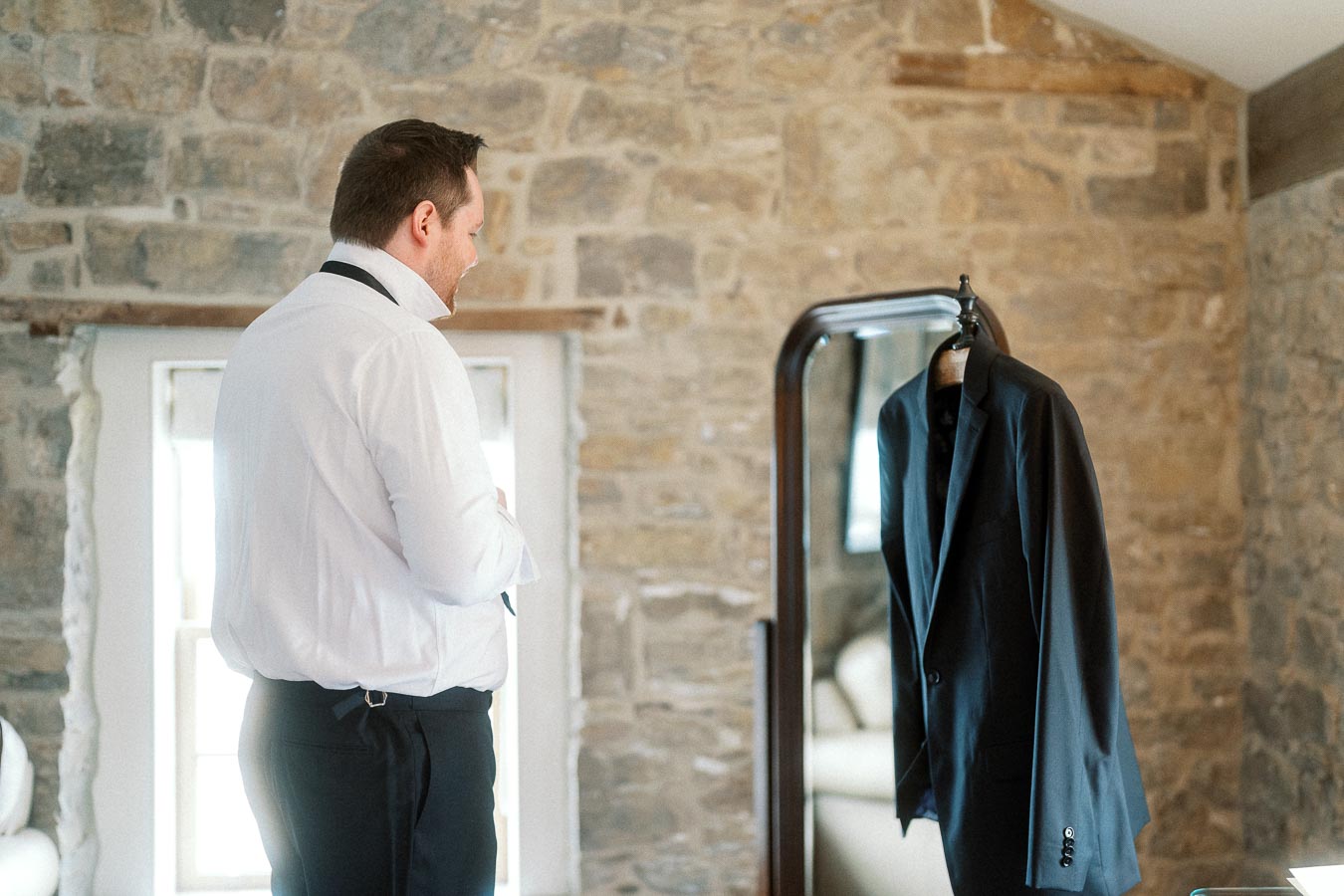 Man in a white dress shirt stands near a hanging suit jacket in a stone-walled room.