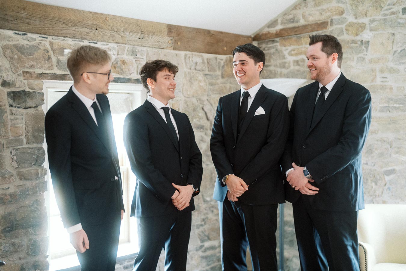 Four men in black suits smiling together in a stone-walled room