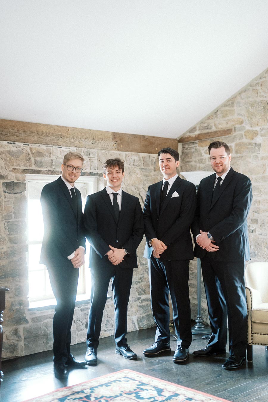 Four men in black suits posing indoors against a stone wall.
