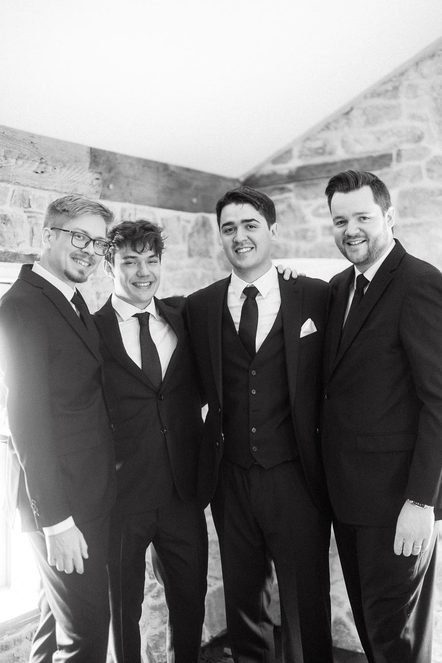 Four men in black suits posing together indoors by a stone wall.