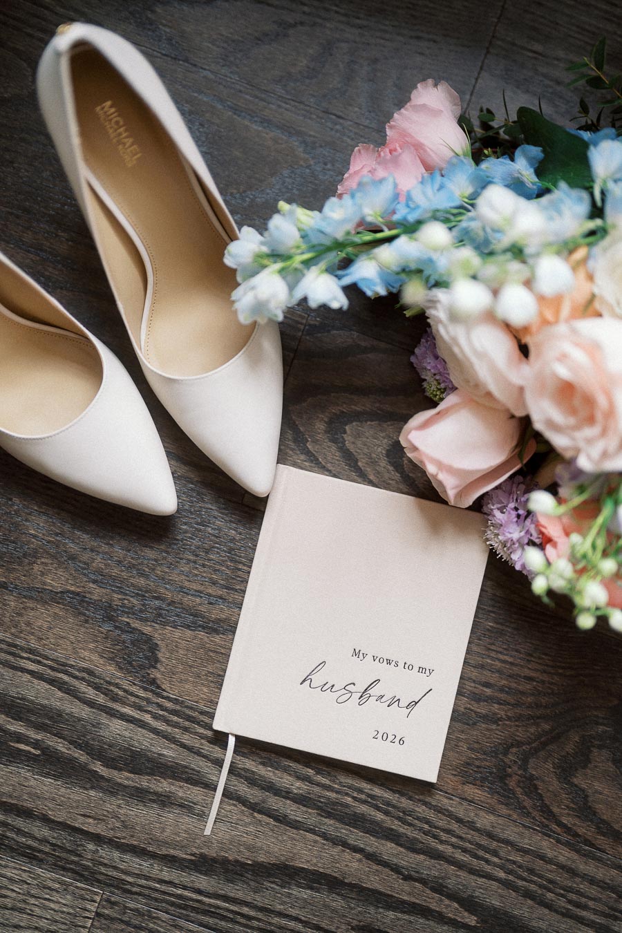 White heels, pastel bouquet, and vow book arranged on dark wood flooring