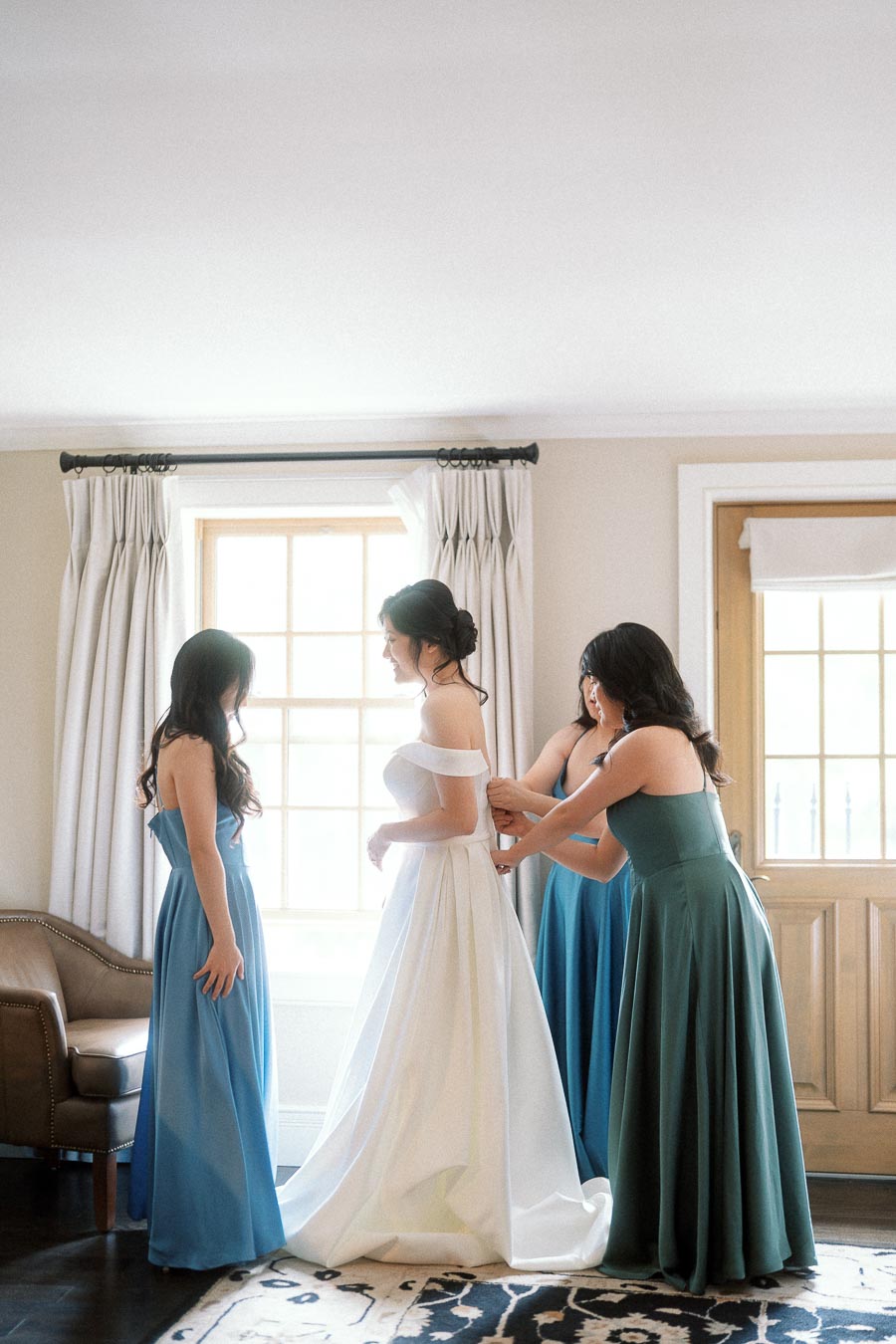 Bride in a white gown being helped by bridesmaids in blue and green dresses