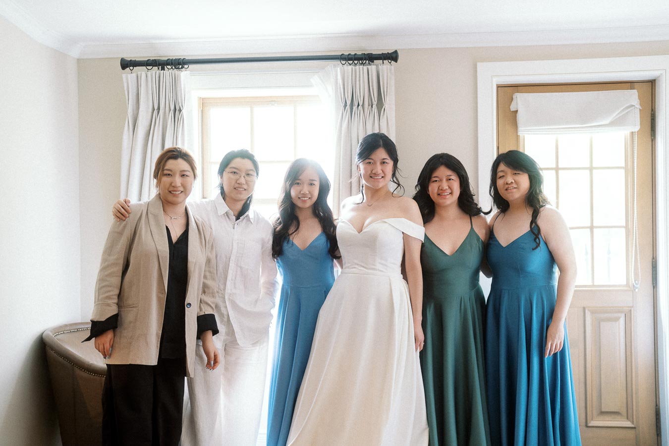 Bride in a white gown posing indoors with five attendants by bright windows