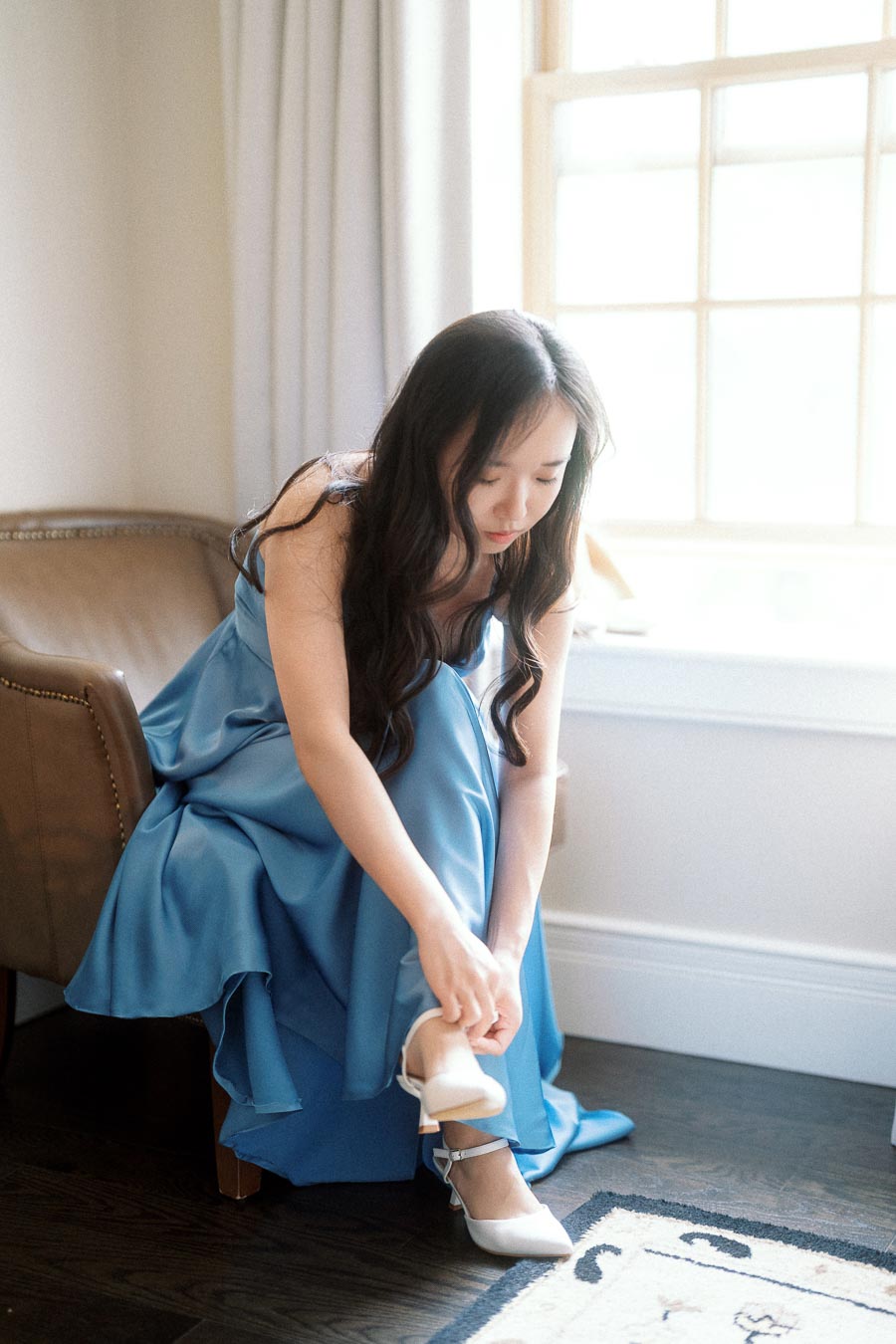 Woman in a blue dress fastening a white heel beside a bright window
