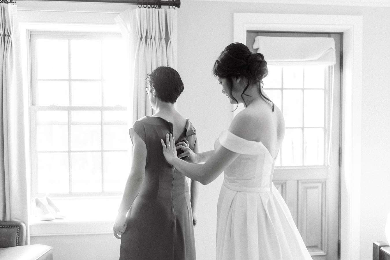 Bride in an off-shoulder gown helps another woman zip her dress by a window.