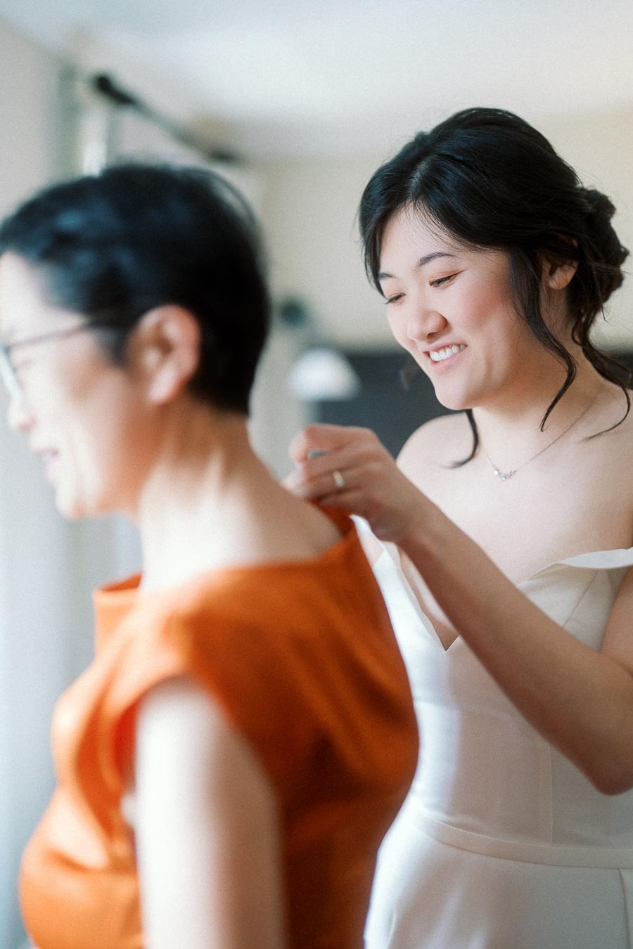 Woman in a white dress fastening a necklace on another woman indoors
