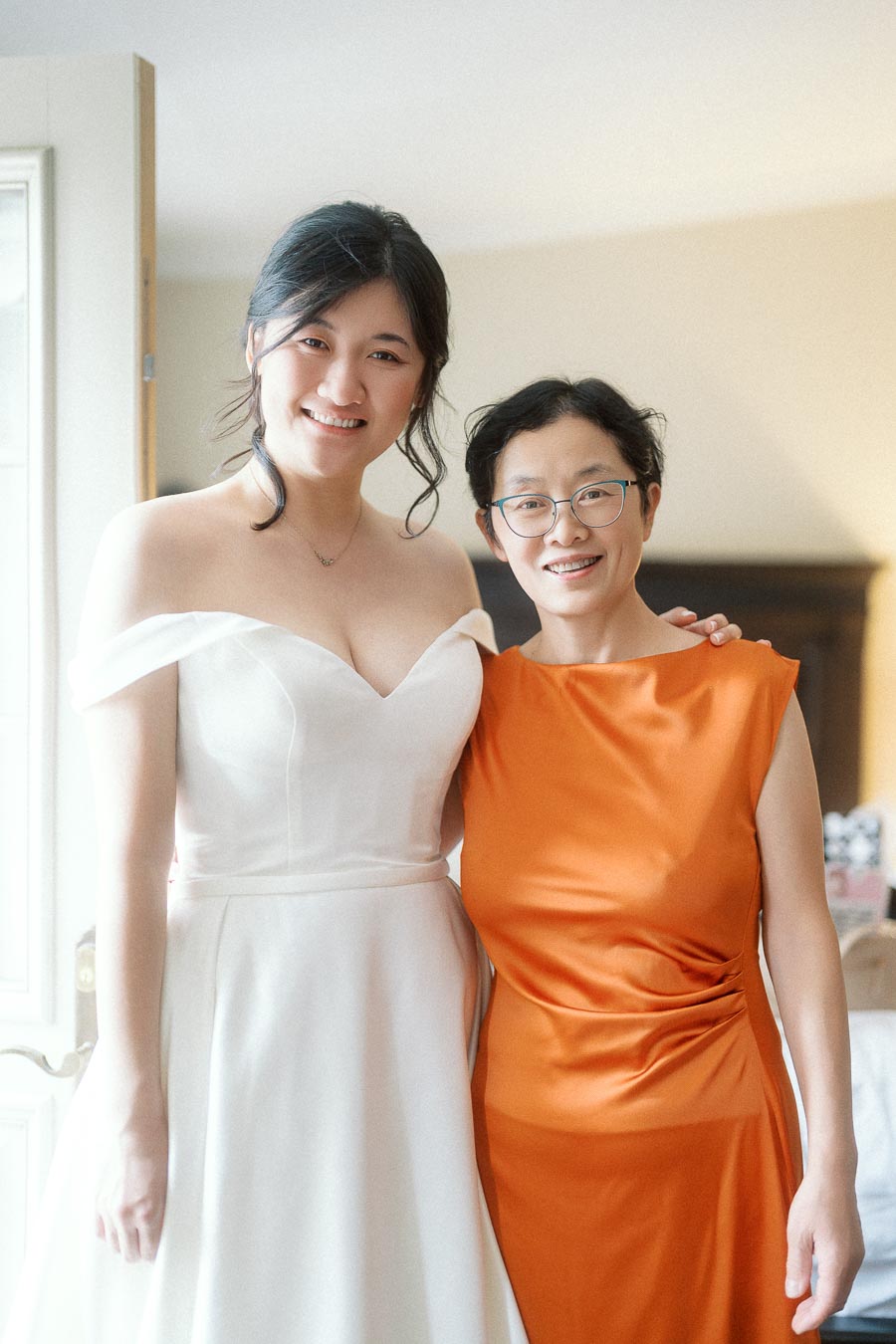 Two women smiling indoors, one in a white gown and one in an orange dress.