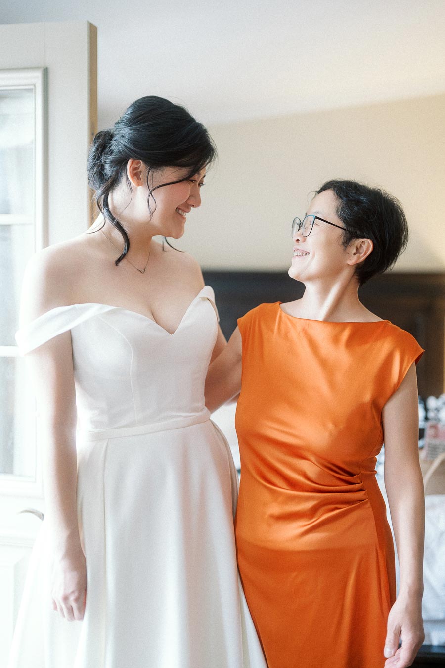 Two women smiling together indoors, one wearing a white off-shoulder gown.