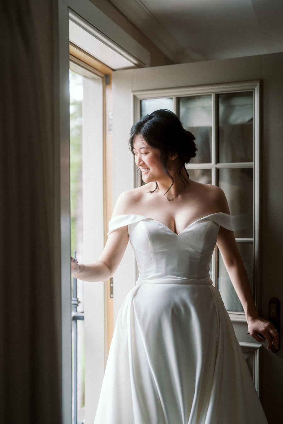 Bride in an off-the-shoulder white gown smiling by an open doorway