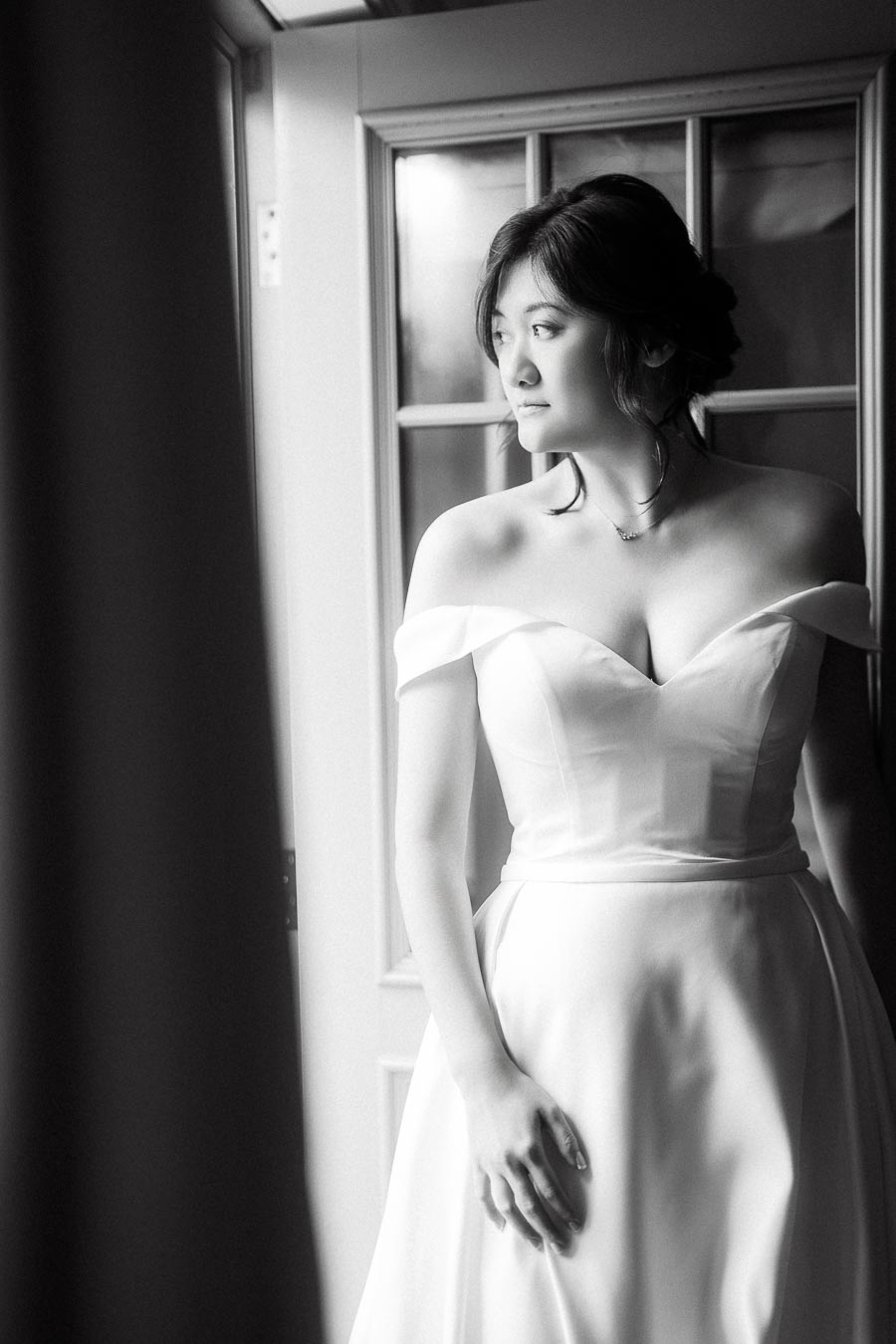 Woman in an off-shoulder white dress standing by a window in black and white