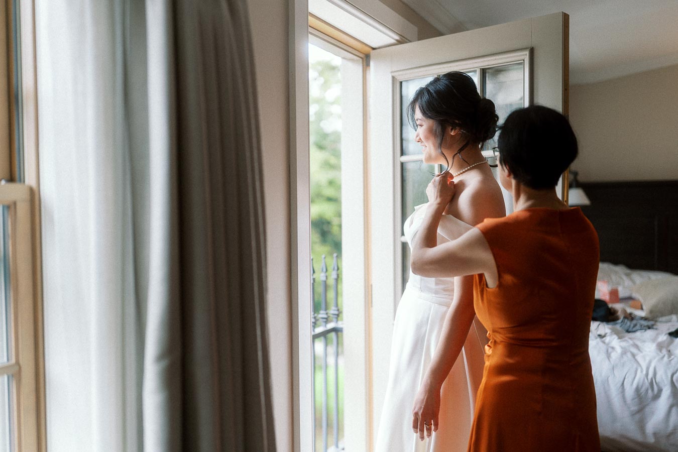 Woman in a white dress has her necklace adjusted beside an open balcony door.